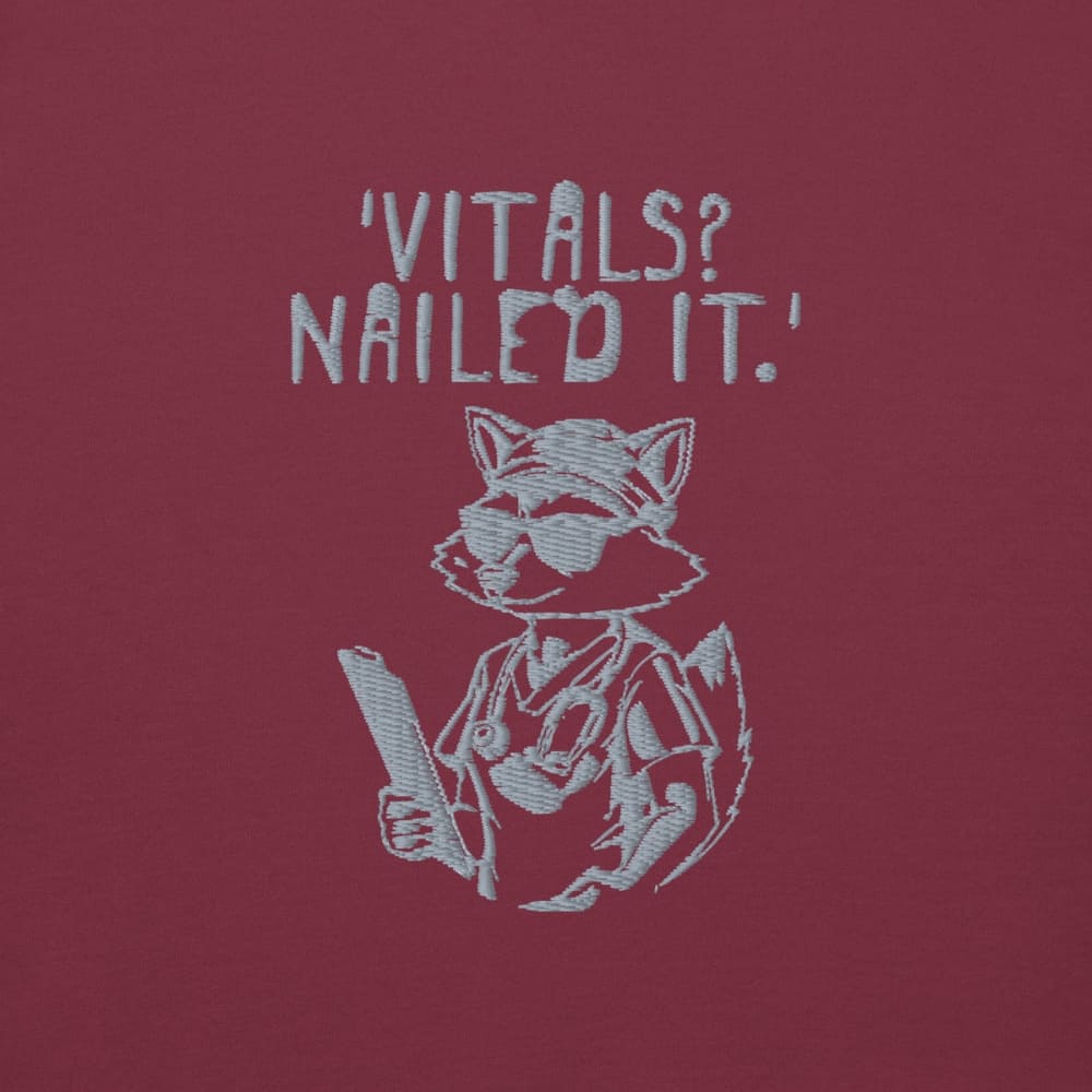Raccoon nurse graphic wearing scrubs and sunglasses holding a clipboard under the quote “Vitals? Nailed It.” – printed on a black Cotton Heritage hoodie