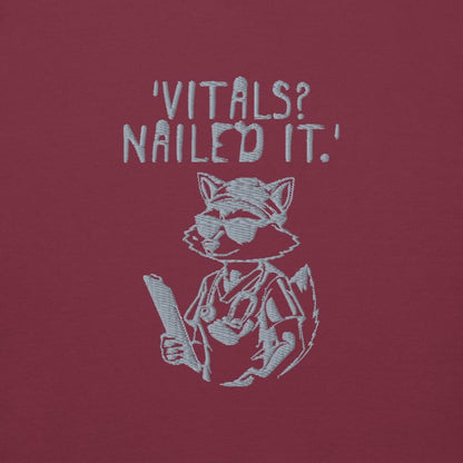 Raccoon nurse graphic wearing scrubs and sunglasses holding a clipboard under the quote “Vitals? Nailed It.” – printed on a black Cotton Heritage hoodie