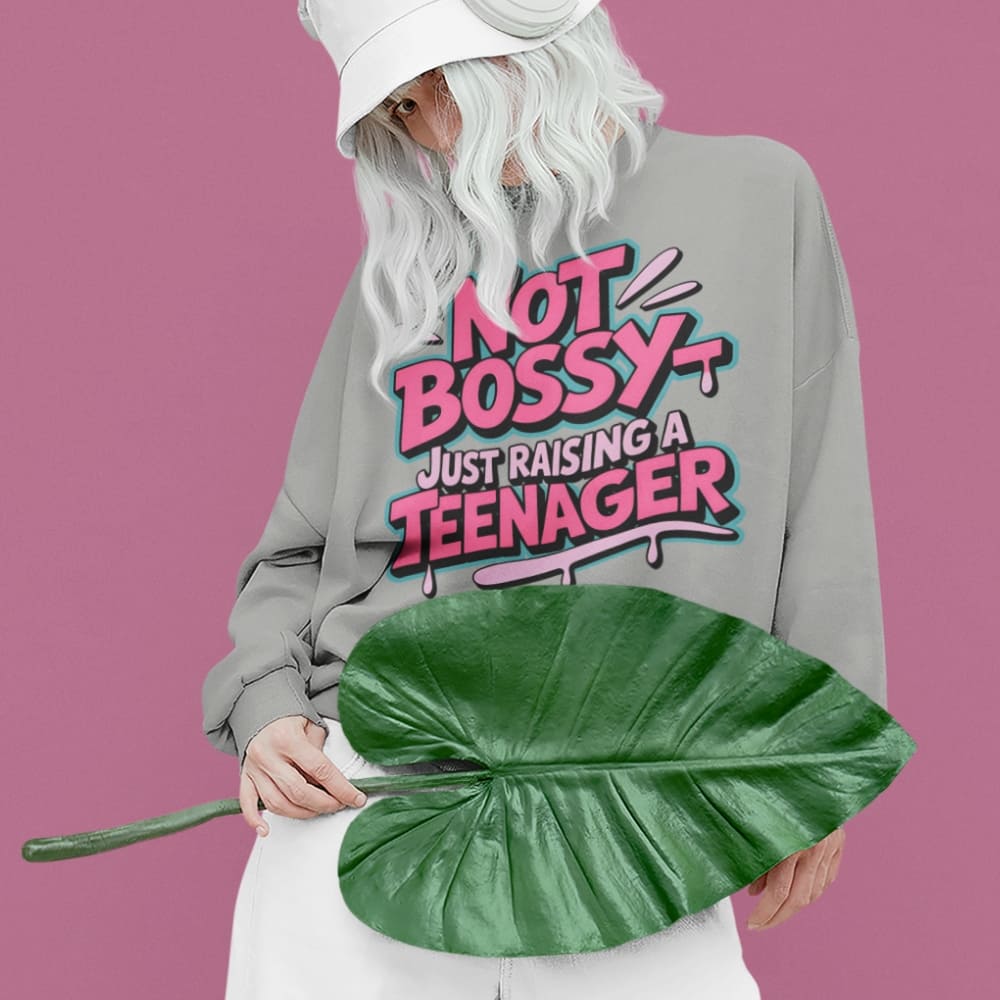 Person wearing a sweatshirt with 'Not Bossy Just Raising a Teenager' text, holding a large green leaf against a pink background.