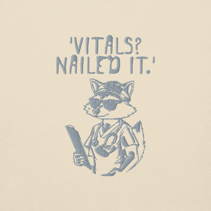 Raccoon nurse graphic wearing scrubs and sunglasses holding a clipboard under the quote “Vitals? Nailed It.” – printed on a black Cotton Heritage hoodie