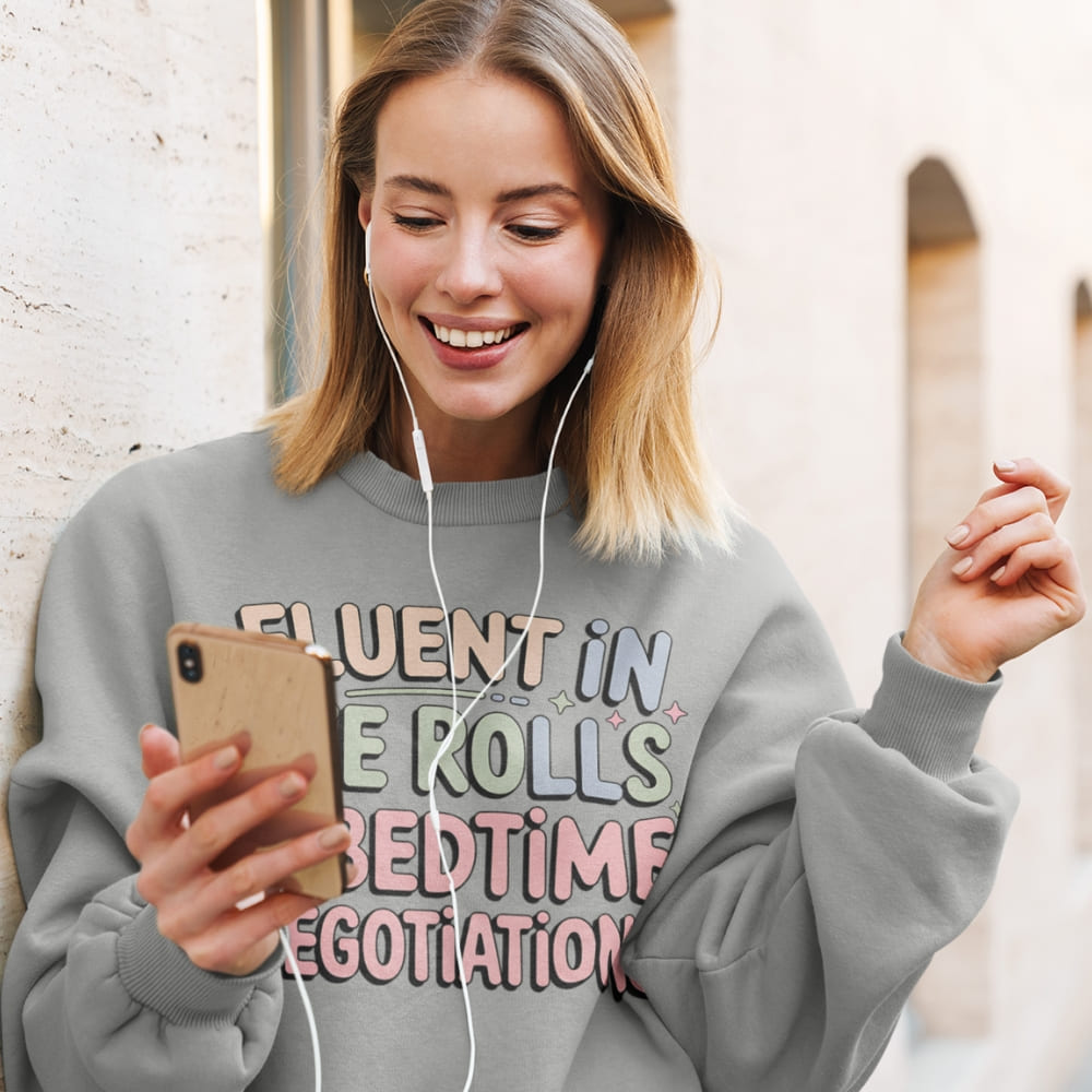Woman wearing a gray sweatshirt with humorous text, using a phone and earphones outdoors.