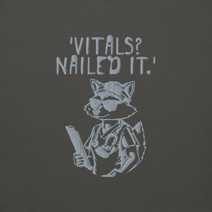 Raccoon nurse graphic wearing scrubs and sunglasses holding a clipboard under the quote “Vitals? Nailed It.” – printed on a black Cotton Heritage hoodie
