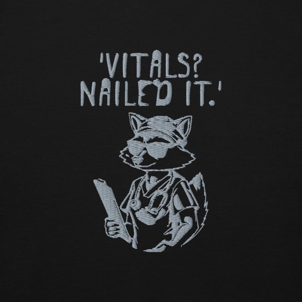 Raccoon nurse graphic wearing scrubs and sunglasses holding a clipboard under the quote “Vitals? Nailed It.” – printed on a black Cotton Heritage hoodie