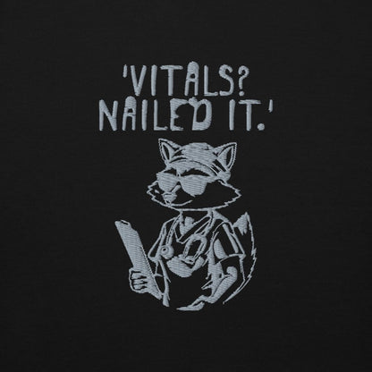Raccoon nurse graphic wearing scrubs and sunglasses holding a clipboard under the quote “Vitals? Nailed It.” – printed on a black Cotton Heritage hoodie