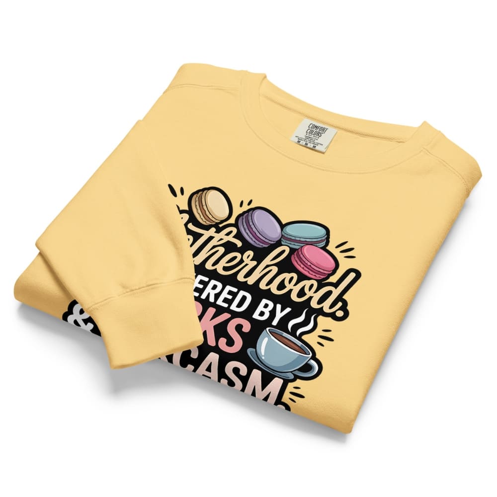 Sweatshirt design with text “Motherhood. Powered by Snacks & Sarcasm.” featuring colorful macarons and a steaming coffee cup on vintage-washed cotton fleece.