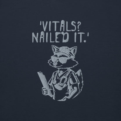 Raccoon nurse graphic wearing scrubs and sunglasses holding a clipboard under the quote “Vitals? Nailed It.” – printed on a black Cotton Heritage hoodie