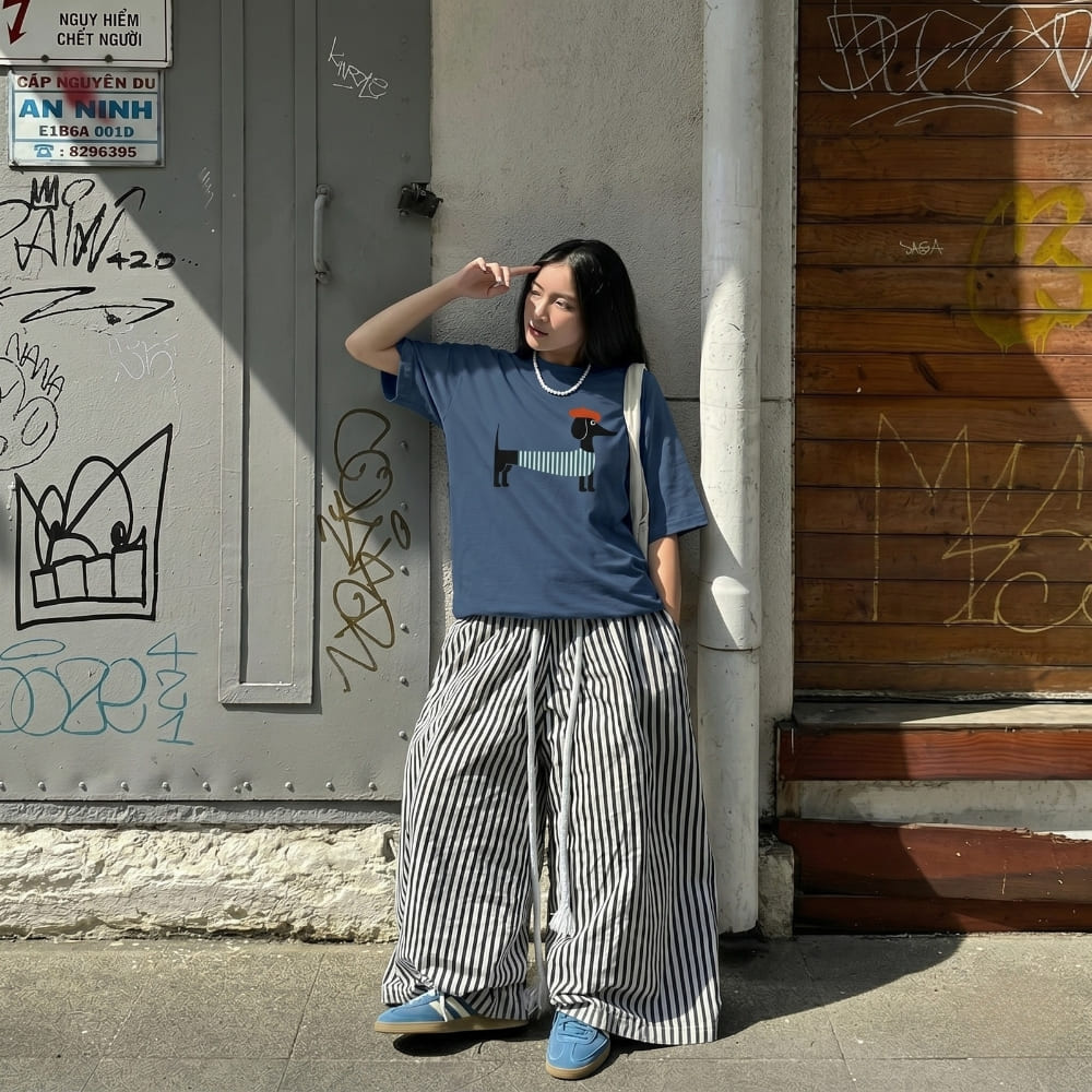 Woman wearing a blue t-shirt with a graphic design and striped pants standing against a graffiti-covered wall.