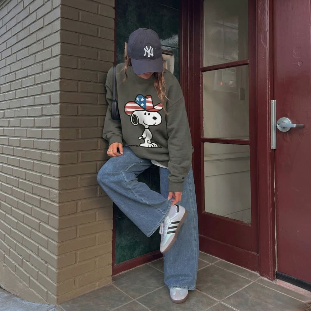 Person wearing a gray sweatshirt with a Snoopy design, blue jeans, and white sneakers, leaning against a brick wall.