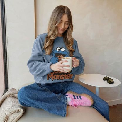 Woman wearing a Cookie Monster sweatshirt, sitting on a couch holding a mug.