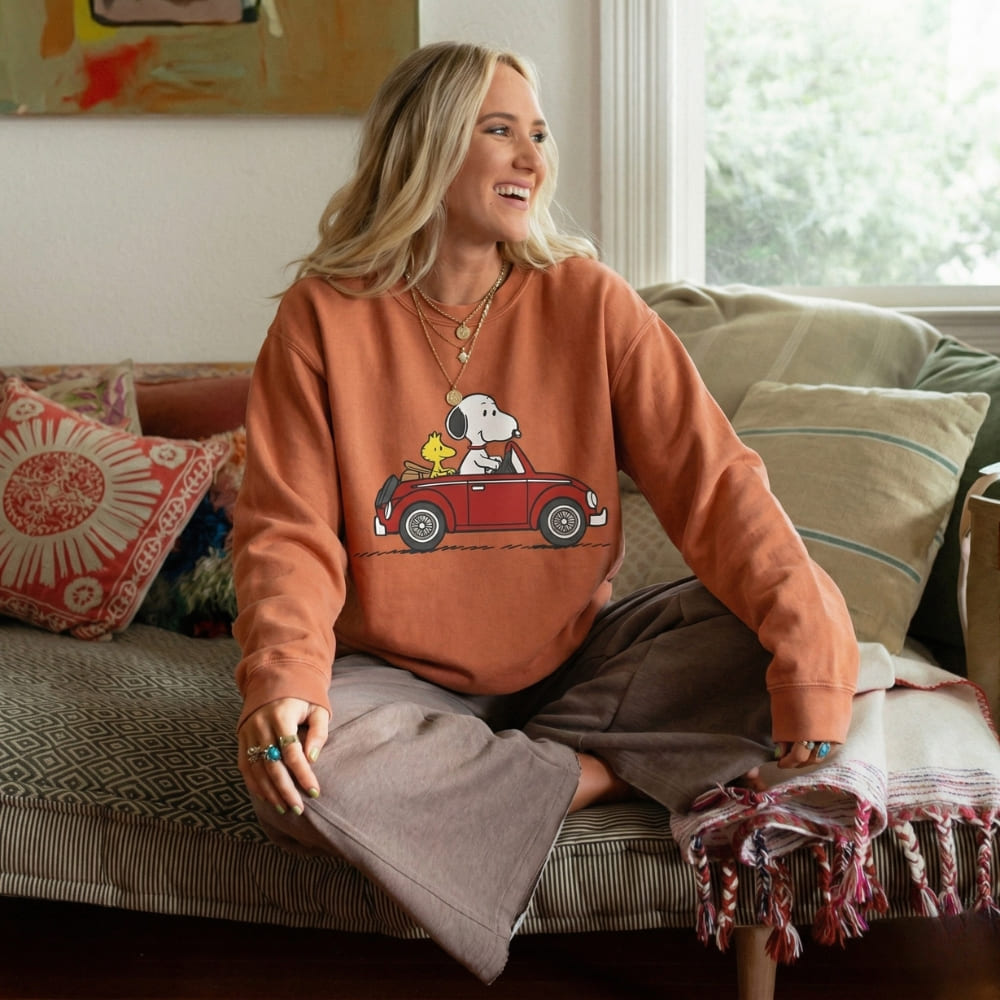 Woman wearing a orange sweatshirt with a graphic design of a dog in a car, sitting on a couch.