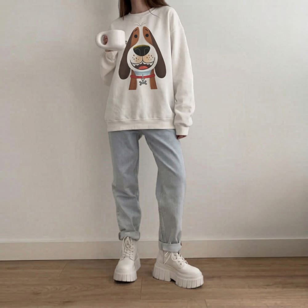 Person wearing a white sweatshirt with a dog graphic, light blue jeans, and white sneakers, holding a white mug.