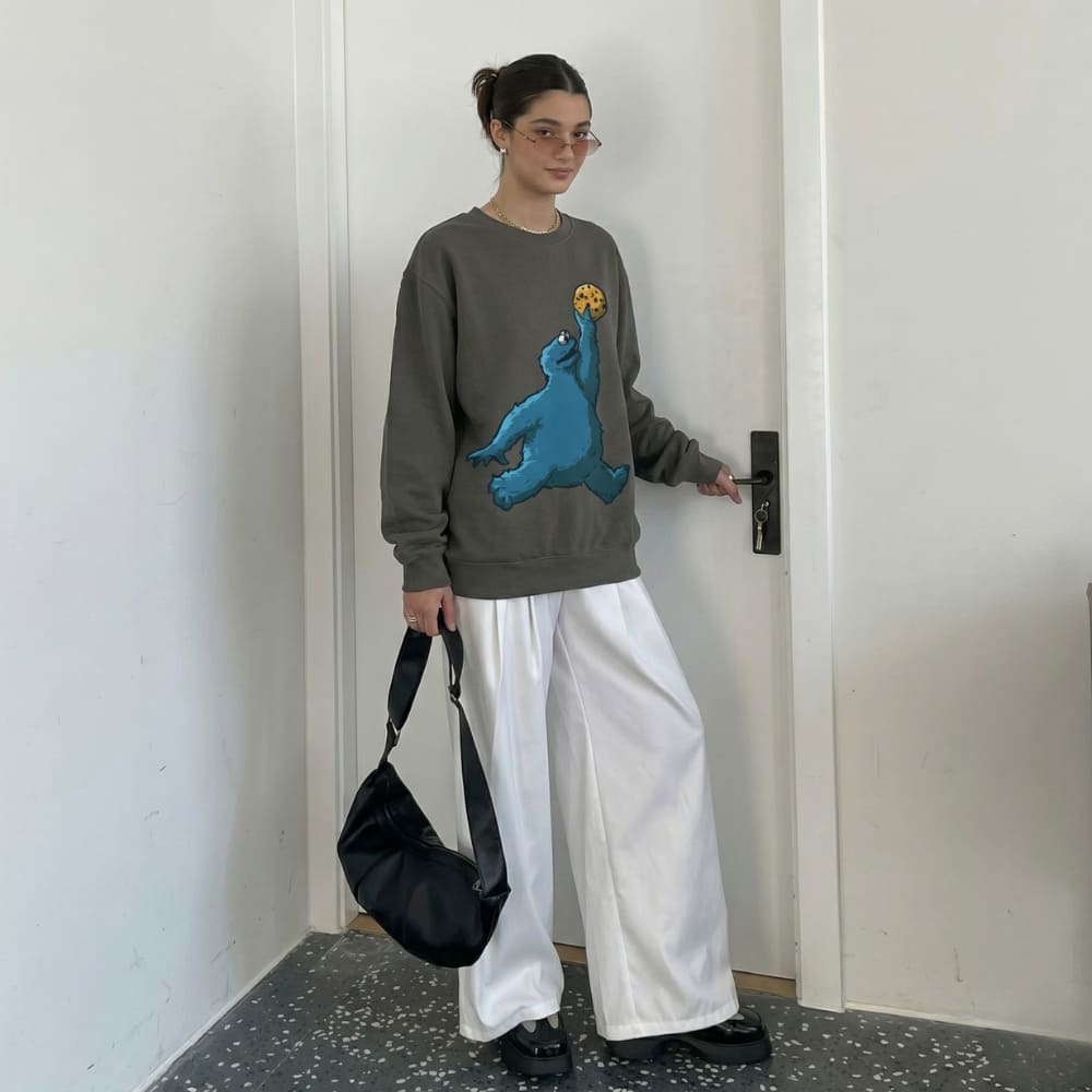Person wearing a gray sweatshirt with a blue graphic design, holding a black bag, standing in a room.