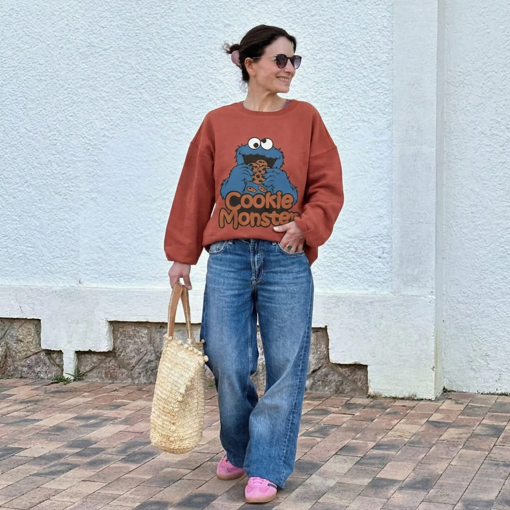 Woman wearing a red sweater with a Cookie Monster design, blue jeans, and pink shoes, standing against a white wall.