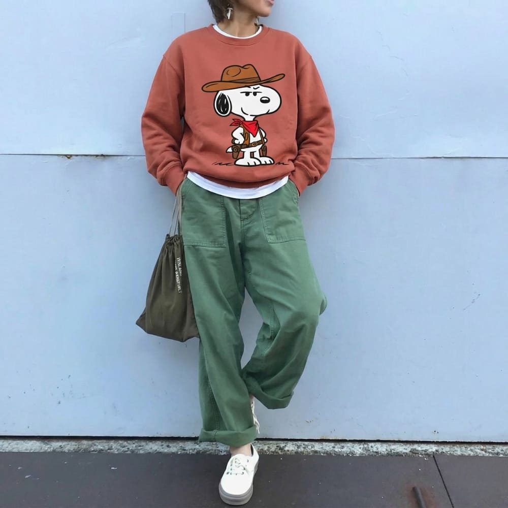 Person wearing a red sweatshirt with a cartoon character, green pants, and white shoes against a light blue wall.