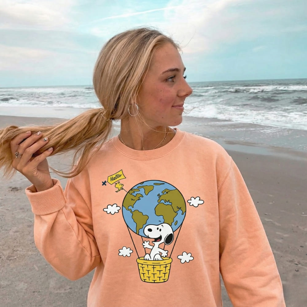 Person wearing a peach-colored sweatshirt with a Snoopy and地球 design on a beach