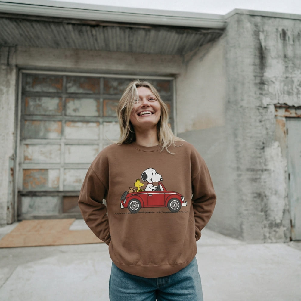 Person wearing a brown sweatshirt with a graphic design of Snoopy and Woodstock in a car, standing in front of an old building.