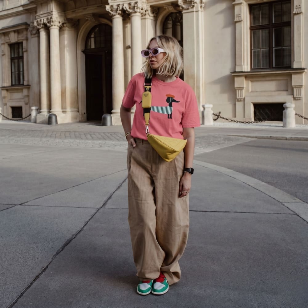 Person wearing a pink shirt with a dog design, beige pants, and a yellow bag standing in front of a classical building.