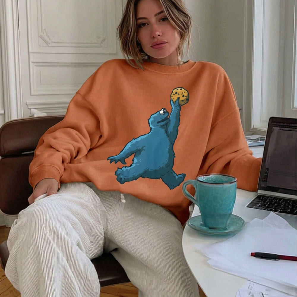 Person wearing an orange sweatshirt with a cookie monster graphic, sitting at a desk with a laptop and mug.