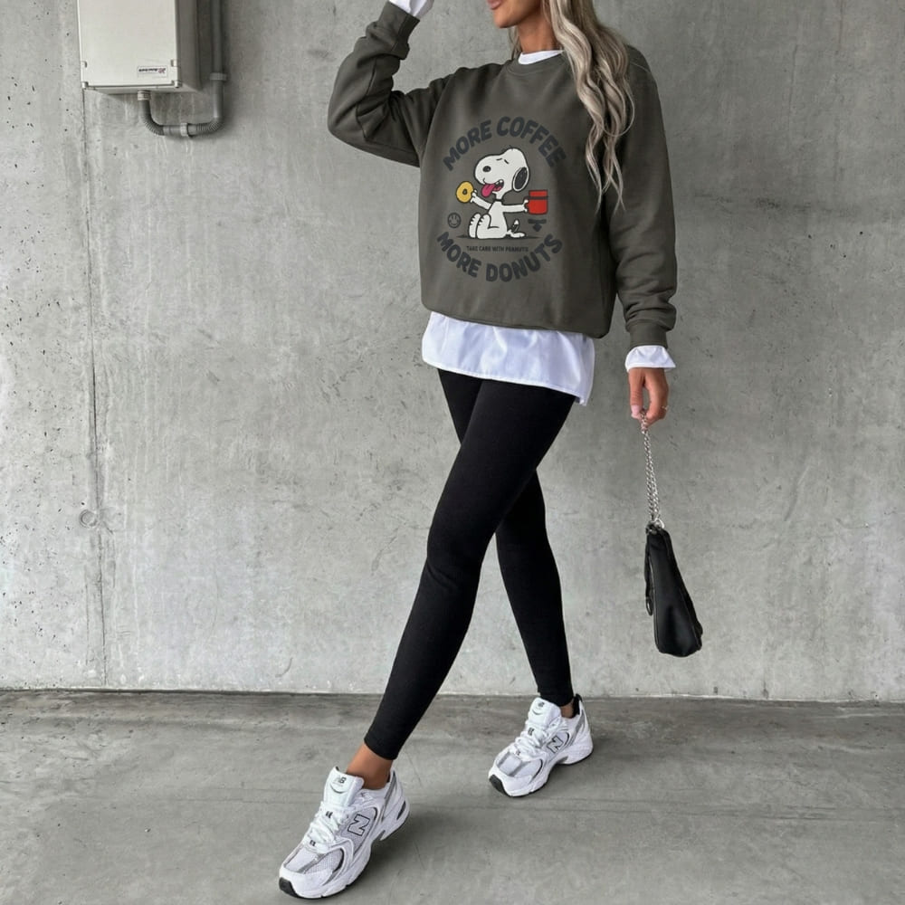 Person wearing a graphic gray sweatshirt with text and a dog design, black leggings, white sneakers, and holding a black handbag against a concrete wall.