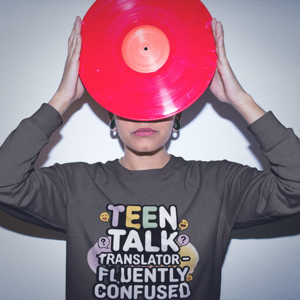 Person wearing a sweatshirt with 'Teen Talk Translator Fluently Confused' text, holding a red vinyl record over their face.