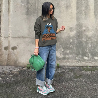 Person wearing a Cookie Monster sweatshirt with a green bag against a concrete wall.