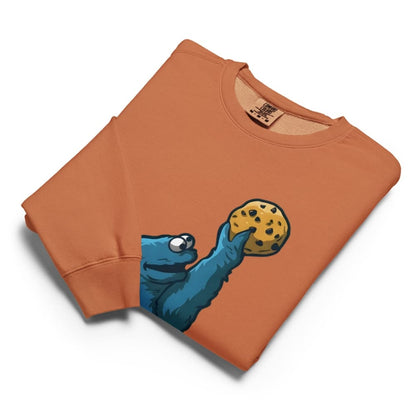 Folded orange sweatshirt with a blue creature holding a cookie graphic on a white background