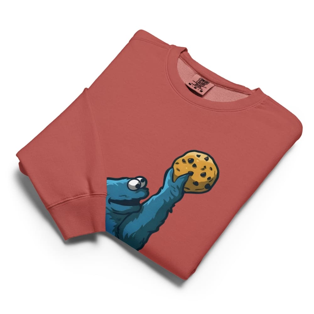 Folded red sweatshirt with a blue creature holding a cookie graphic on a white background