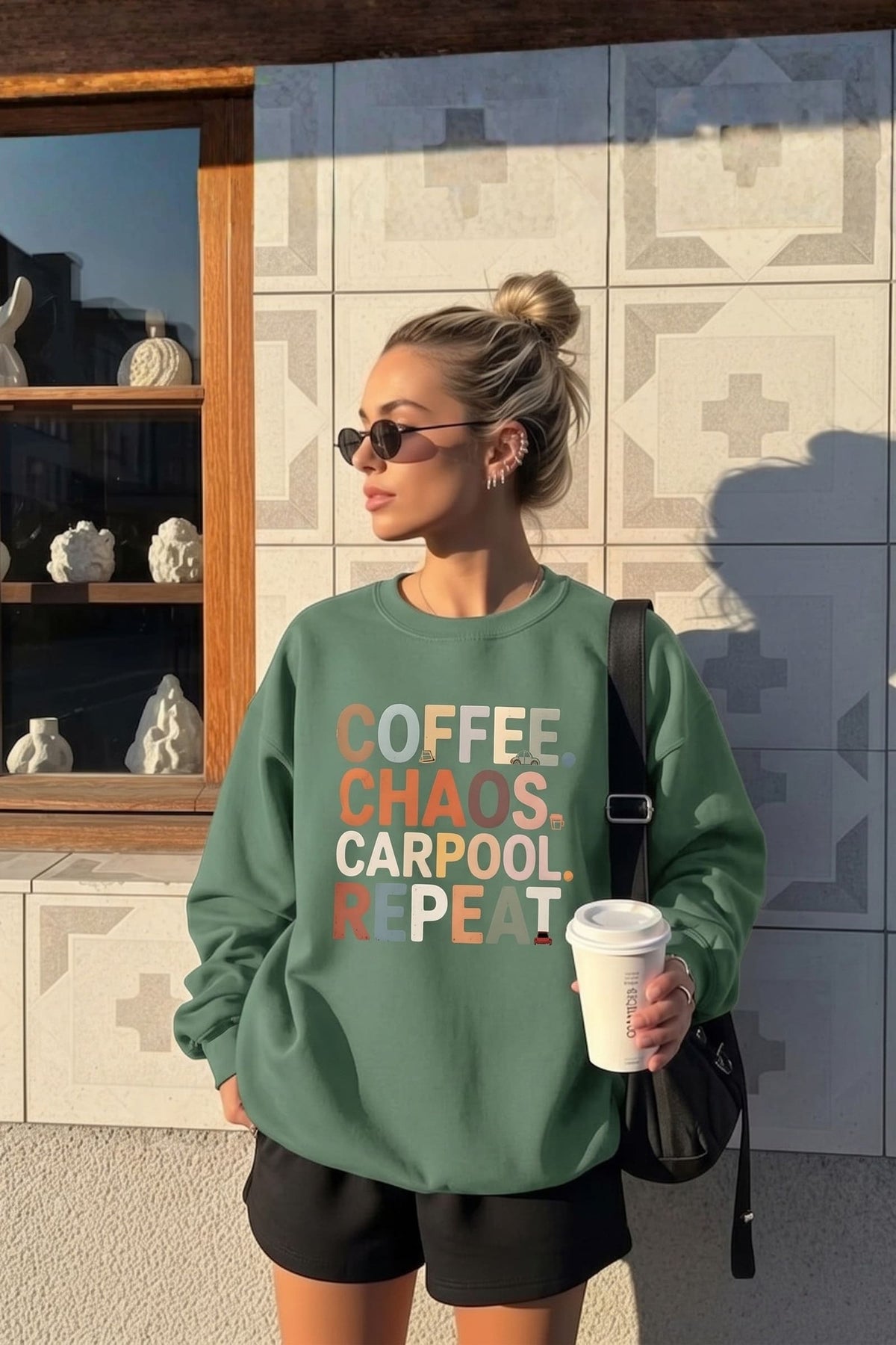 Woman in sunglasses wearing the Coffee Chaos Carpool™ sweatshirt in green, holding a takeaway coffee outside a sunlit tiled storefront